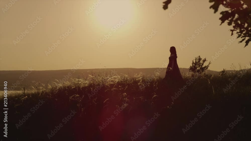 Alone medieval peasant girl is walking on field in background of red sunset sky. She is moving to an edge of ground, out from forest