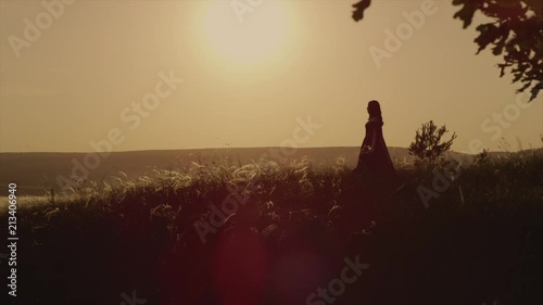 Alone medieval peasant girl is walking on field in background of red sunset sky. She is moving to an edge of ground, out from forest