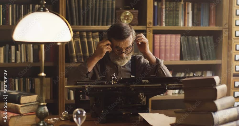 Portrait shot of the old gray haired man putting on glasses before working on the typewriter in the library cabinet.