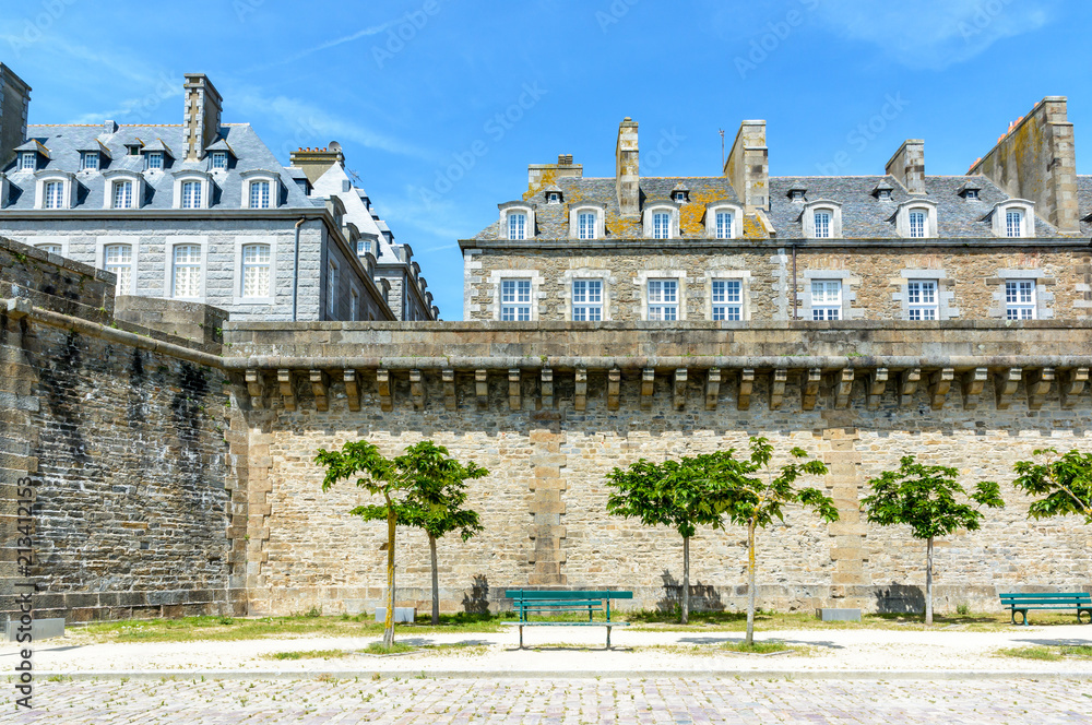 View of the surrounding wall of the city of Saint-Malo in Brittany, France, a small park with trees and benches at the foot of the wall and granite residential buildings sticking out above the rampart