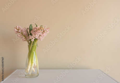 Close up of soft pink hyacinths in glass vase on white table against beige wall (selective focus) © Natalie Board