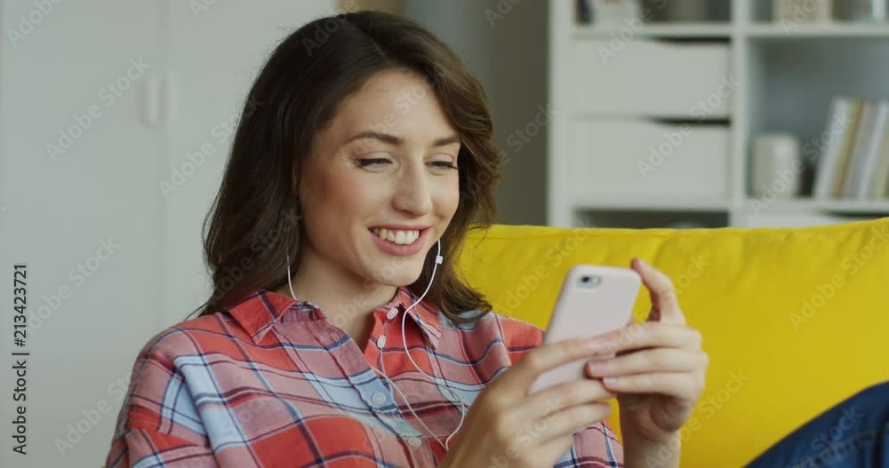 Portrait of the young smiled Caucasian woman in the headphones taping and scrolling on her smartphone at home. Portrait of the young smiled Caucasian woman in the headphones taping and scrolling on her smartphone at home.