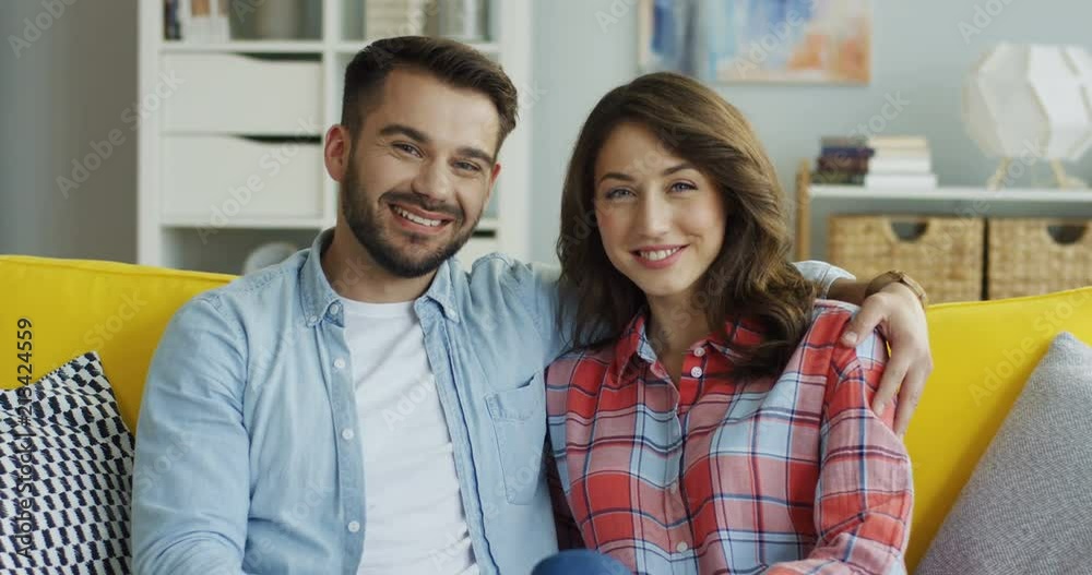 Portrait shot of the young Caucasian smiled boyfriend and girlfriend sitting on the yellow sofa and posing to the camera at home. Portrait shot of the young Caucasian smiled boyfriend and girlfriend sitting on the yellow sofa and posing to the camera at home.