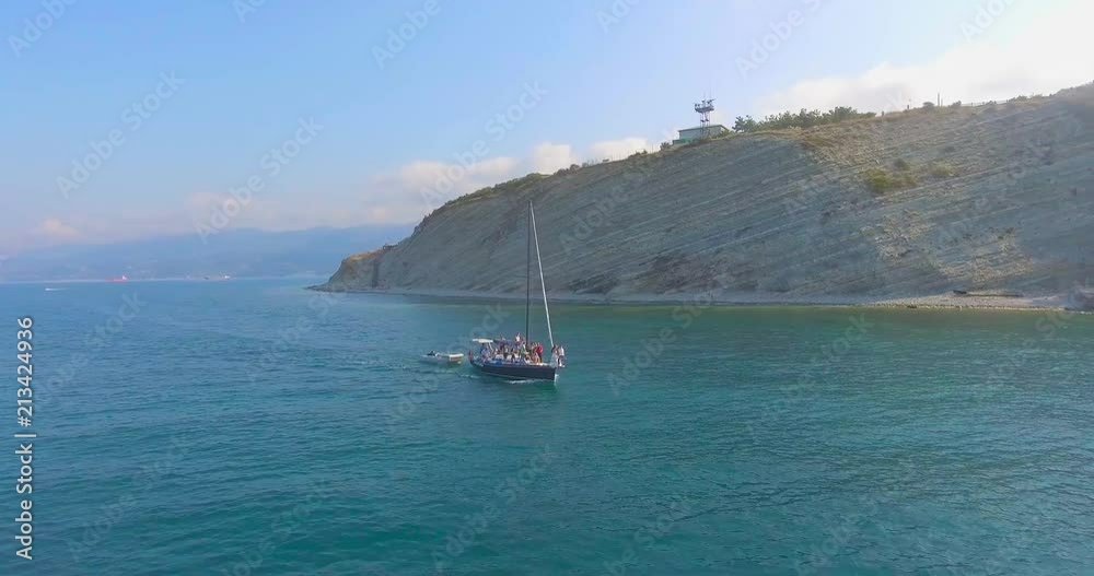 Amazing aerial motion around white yacht floating quickly on azure surface of sea against huge gray rock under boundless blue sky Amazing aerial motion around white yacht floating quickly on azure surface of sea against huge gray rock under boundless blue sky
