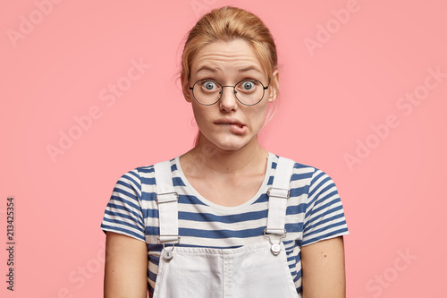 Worried European female bites lower lip, looks with puzzled expression, wears fashionable outfit, stands against pink background. Frustrated embarrassed woman feels nervous while makes decision