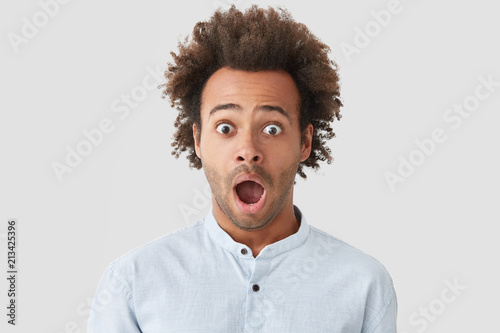 Stunned handsome mixed race young male with bushy dark hair, keeps jaw dropped, opens eyes widely, can`t believe in bad fortune, expresses great surprisement, isolated over white background.
