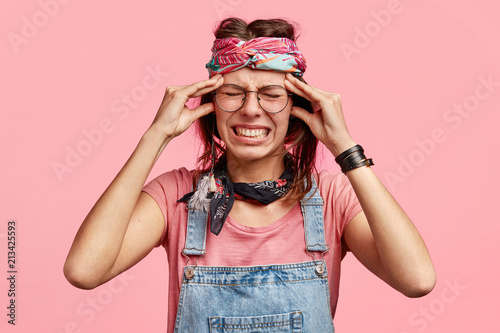 Fényképezés Studio shot of desperate young hippie woman has terrible headache, clenches teeth, has sorrowful expression, has problems, going to cry, wears stylish headband, belongs to specific subluture