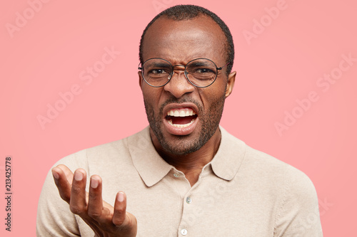 Displeased dark skinned guy frowns face in discontent, feels aversion, shows teeth, stands indoor against pink wall, dressed in beige t shirt. African American man under pressure. Anger and annoyance