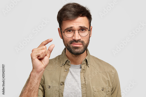 That`s all I have! Displeased bearded young male model shapes something tiny with hand, makes small gesture, discontet with salary, stands alone against white studio wall. Man talks about size