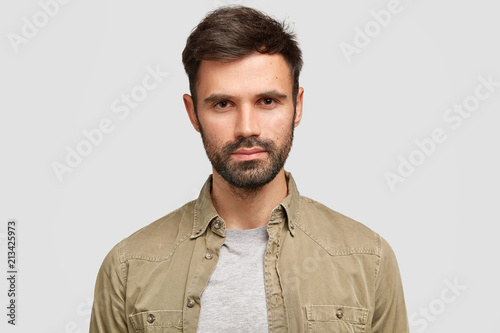 Photo of confident bearded male with dark hair, has serious facial expression, looks directly at camera, thinks about future job, dressed in fashionable shirt, stands against white studio wall