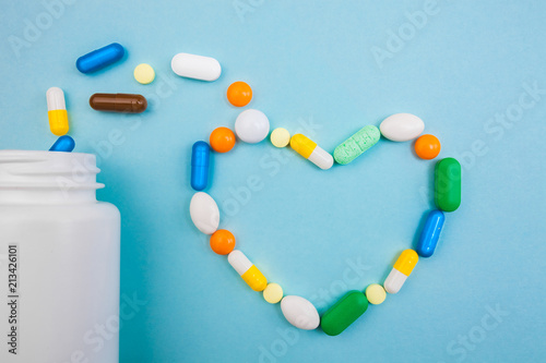 Multi-colored tablets and heart-shaped capsules, white bottle for tablets, pharmaceutical medicine pills on blue background, an analgesic against diseases