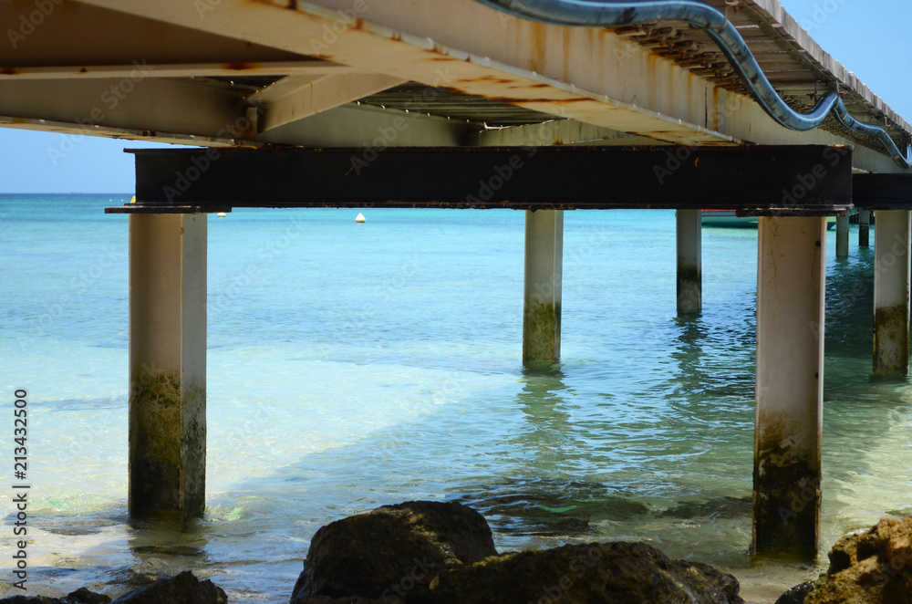 A wooden jetty stretching out into a tropical ocean. Sand and rocks can be seen through the clear water.