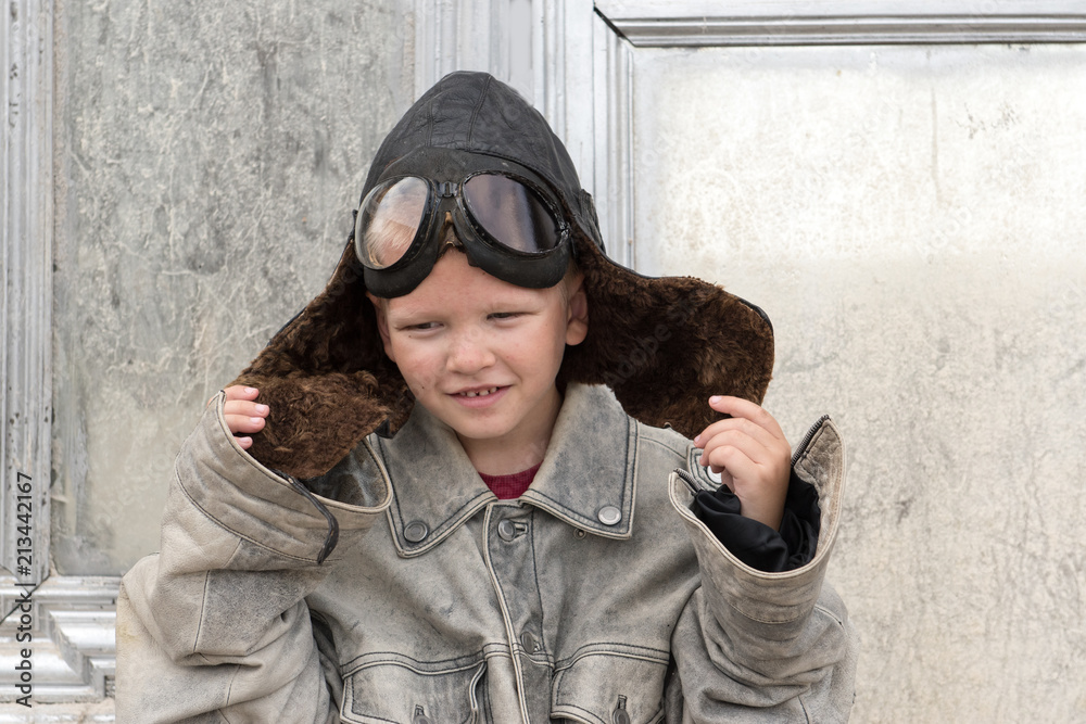 Funny child with independent character wore leather flying hat, pilot's glasses, jacket. Boy dreams of traveling, flying on airplanes, victories, adventures. Concept of man's education, patriotizm Funny child with independent character wore leather flying hat, pilot's glasses, jacket. Boy dreams of traveling, flying on airplanes, victories, adventures. Concept of man's education, patriotizm