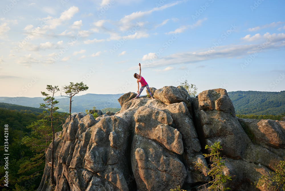 Attractive athletic girl doing complicated yoga exercises on top of huge pile of rocks on green mountains and clear blue sky background. Climbing, tourism, fitness and healthy lifestyle concept.