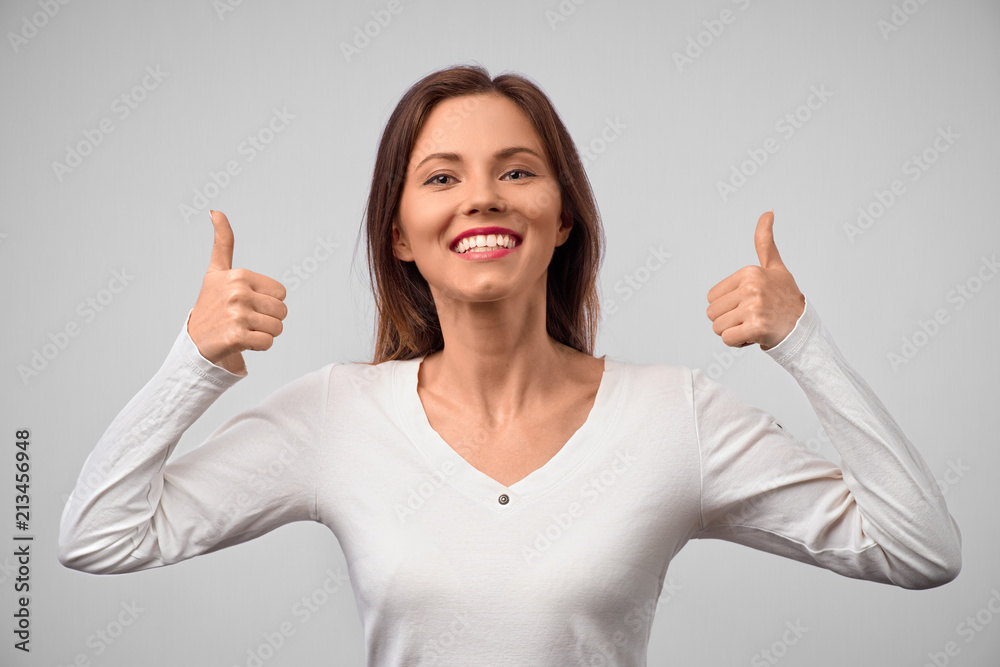 Happy young caucasian female wearing white long sleeved shirt making thumb up sign and smiling cheerfully, showing her support and respect to someone. Body language. I like that. Good job