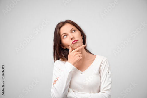 Close up portrait of pretty young woman thinking about problem, confused emotion, isolated, wearing white shirt, hipster style, dark hair, holding finger at lips, looking up, having idea
