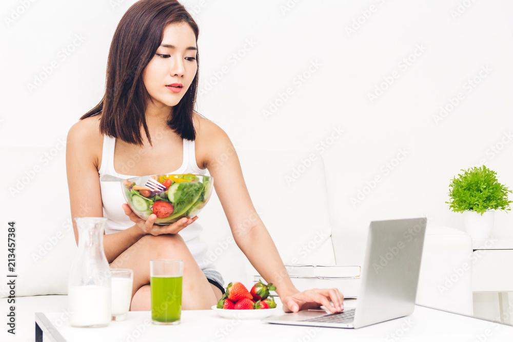 Woman holding healthy fresh salad in a bowl and working on a laptop computer while relaxing on sofa at home.dieting concept.healthy lifestyle with green food