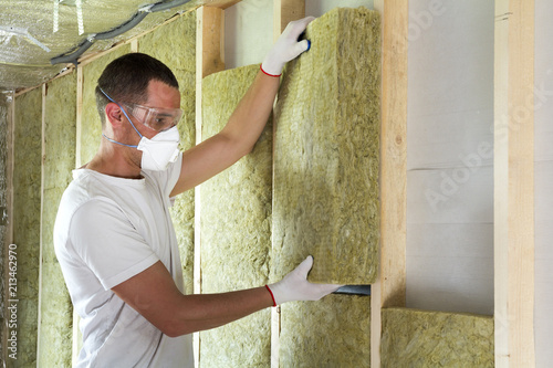 Foto Worker in protective goggles and respirator insulating rock wool insulation in wooden frame for future house walls for cold barrier