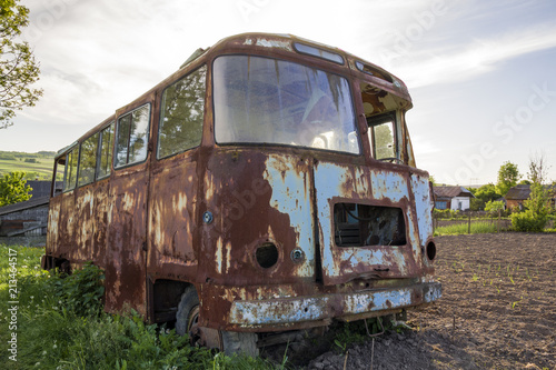 Close-up of old forsaken passenger bus with broken windows rusting in high green weedy grass on edge of plowed brown field on bright spring day under blue morning sky on small peasant huts background.