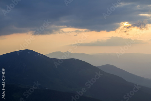 Wide panorama, fantastic view of covered with morning mist green Carpathian mountains at dawn under dark clouds and light pink sky before sunrise. Beauty of nature, tourism and traveling concept.
