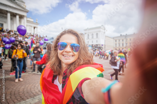 A beautiful young happy woman with a rainbow flag, a symbol of the LGBT community, makes a selfie on a mobile phone on a pride in a European city. Human Rights, Equality, LGBT, Freedom and Happiness