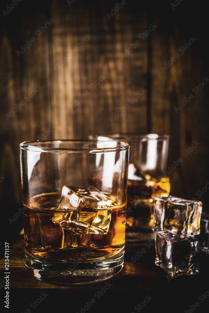 Two whiskey shot glasses on dark wooden background, with ice cubes, copy space