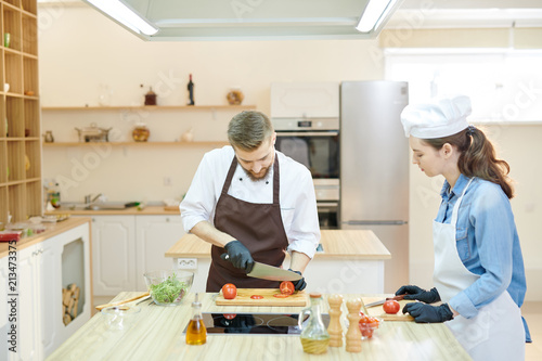 Portrait of professional cook working in restaurant kitchen with female su-chef, both cutting vegetables standing at wooden workstation, copy space