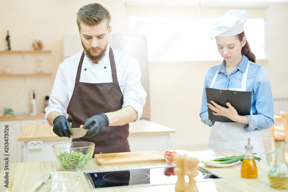 Waist up portrait of two professional chefs working in restaurant kitchen together cooking and seasoning salad standing at wooden table Waist up portrait of two professional chefs working in restaurant kitchen together cooking and seasoning salad standing at wooden table