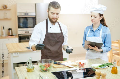 Portrait of two young professional chefs working in restaurant kitchen together cooking and seasoning salad standing at wooden table