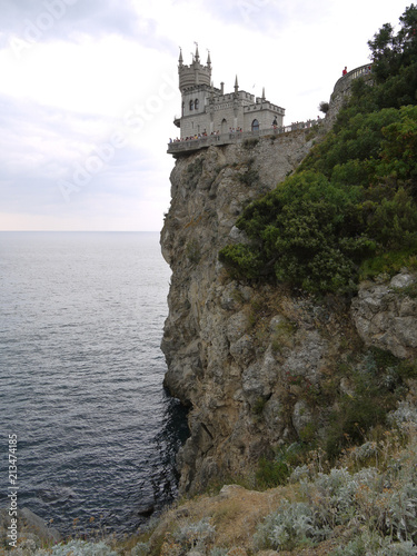 A castle with turrets located at the very edge of a cliff standing in the sea. With fences around the edges and tourists looking at local beauty from a bird s eye view.