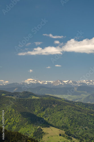 Mountains covered with forest and meadows, snowy mountains and blue sky with clouds. Scenic landscape shot taken at sunny spring day. Concept of clear environment. Natural background.