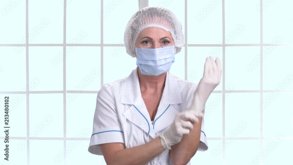 Middle aged female doctor or nurse puts on gloves. Mature woman surgeon in protective mask and cap putting on white protective gloves indoor in hospital, checkered windows background.
