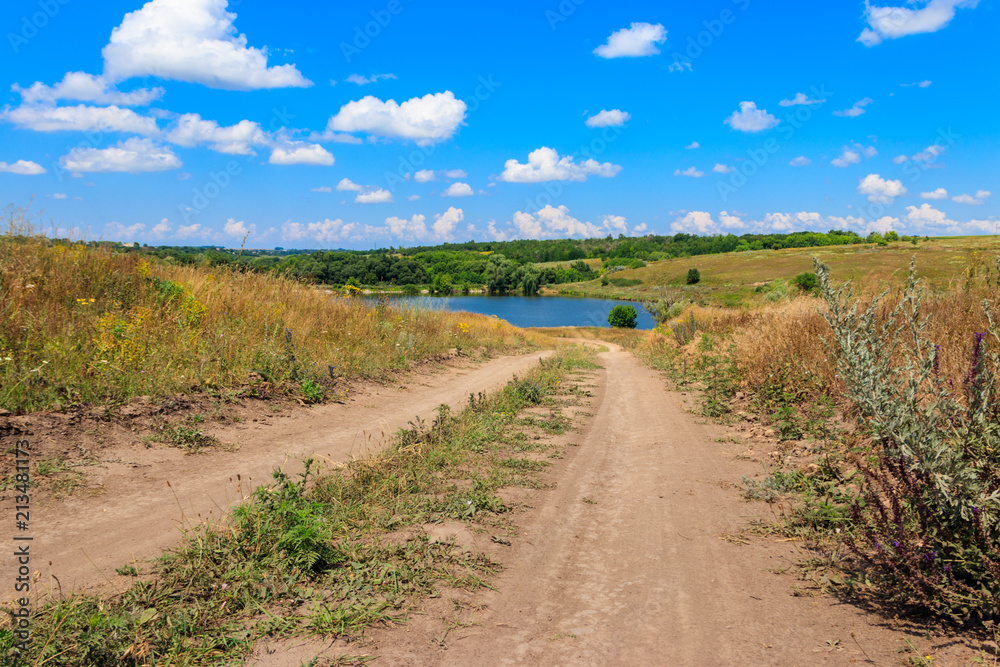 Summer landscape with dirt road to the lake and blue sky with white clouds