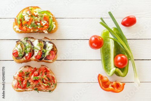 Bruschetta with fresh tomatoes and cheese, eggplant and mozzarella, sweet pepper and goat cheese on white boards with fresh vegetables. Top view. Copy space.