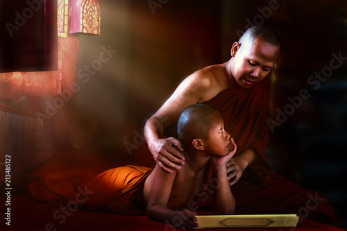 Thai Buddhist monk teaching Buddhist novice Buddhism lessons from Buddist scripture in dark temple with light ray shining from window