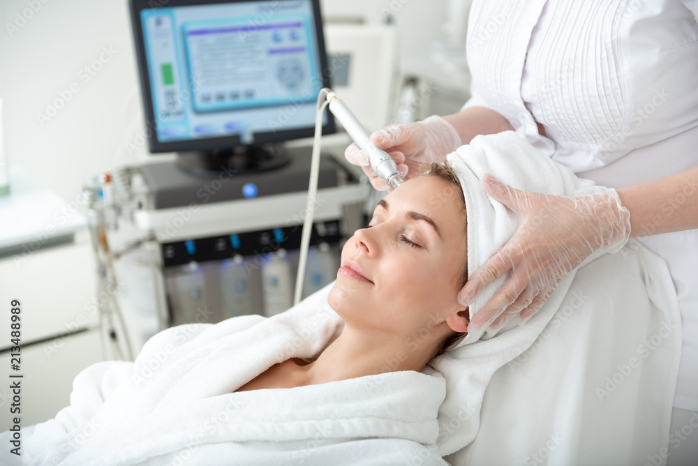Side view beaming lady relaxing on couch while having procedure with special appliance in beauty salon. Health concept Side view beaming lady relaxing on couch while having procedure with special appliance in beauty salon. Health concept