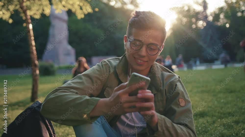 portrait young man with glasses using smartphone listen music on headphone sitting in park smile feel happy sunset sunlight student mobile holding handsome message boy hand caucasian slow motion