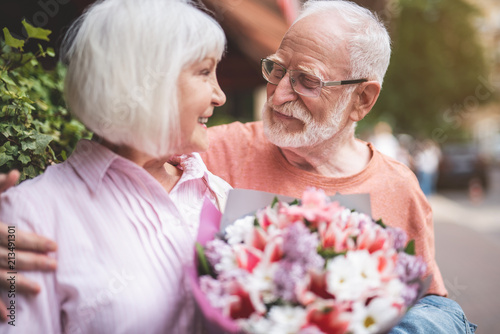 My love. Side view of delighted senior man and woman standing outdoors and smiling. Elderly female is holding beautiful bunch of flowers in hands