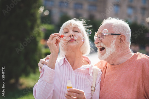 Cheerful lady holding bottle with soap water and blowing bubbles on street. Exited mature male is hugging and looking at her with content. They are embracing each other with pleasure