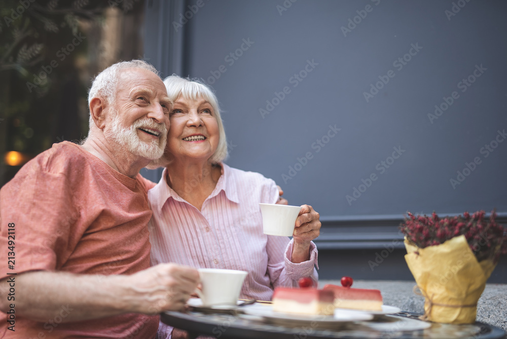 Waist up portrait of smiling couple sitting at table and drinking tea outside. They are hugging and looking sideways with sincere joy Waist up portrait of smiling couple sitting at table and drinking tea outside. They are hugging and looking sideways with sincere joy