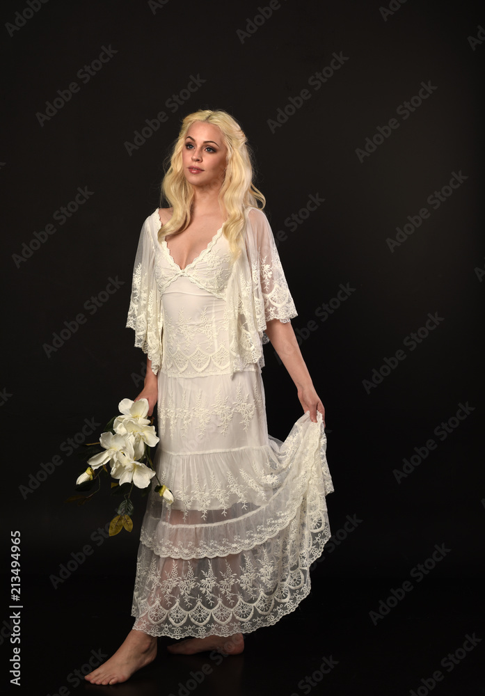 full length portrait of blonde girl wearing white lace dress. standing pose, holding flowers. black studio background. full length portrait of blonde girl wearing white lace dress. standing pose, holding flowers. black studio background.