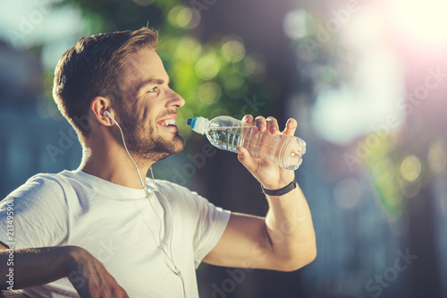 Profile of delighted smiling young athlete holding bottle of water in park. He is thirsty and content after tiring workout