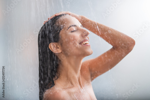 Side view beaming lady locating under stream of water. She holding hair with arms photo