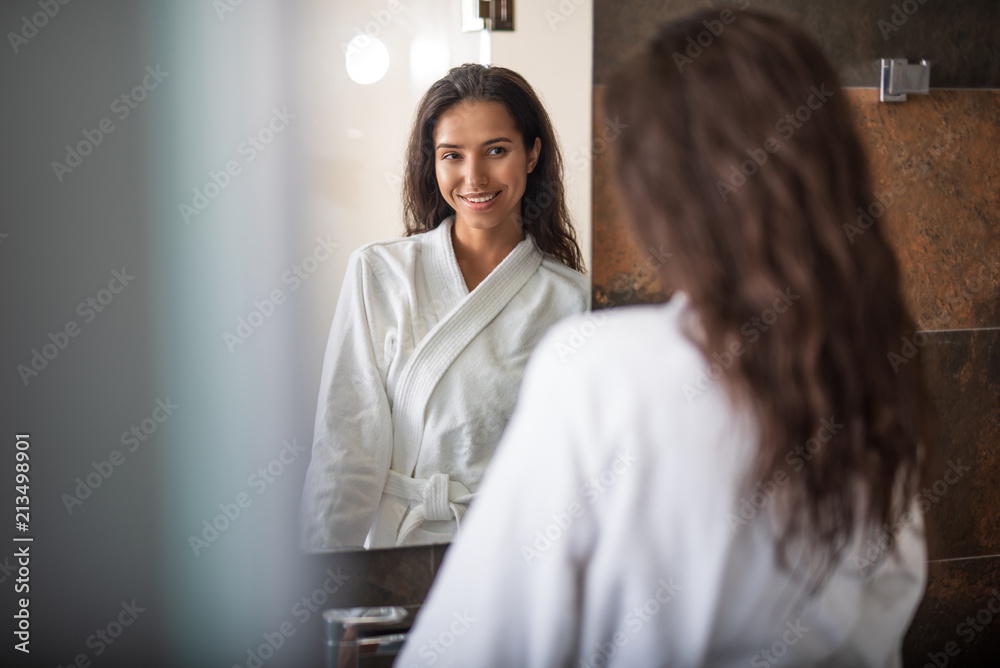 Portrait of beaming pretty girl looking at mirror. She wearing cozy bathrobe while locating in apartment Portrait of beaming pretty girl looking at mirror. She wearing cozy bathrobe while locating in apartment