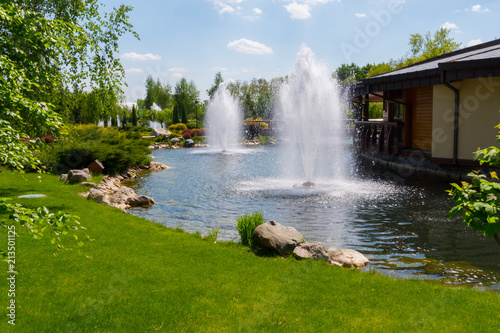 Refreshing transparent fountains in the middle of a pond against a blue sky with a green lawn on one side and a restaurant on the other.