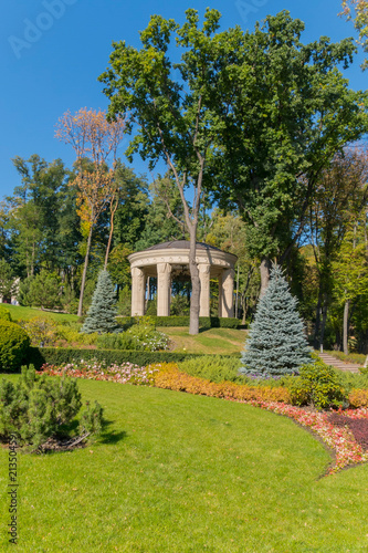 A massive stone pavilion with wide columns and a black roof against a green lawn and tall trees © adamchuk_leo