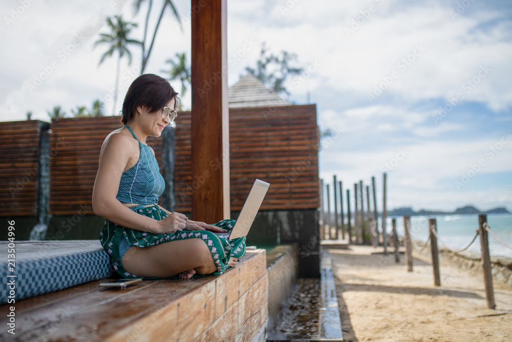 Asian woman working on laptop in pavilion at beachfront in sunny day