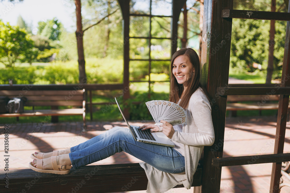 Obraz premium Young successful businesswoman holding bundle of dollars, cash money. Woman working on modern laptop pc computer in city park in street outdoors on nature. Mobile Office. Freelance business concept.