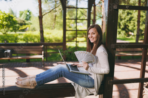 Young successful businesswoman holding bundle of dollars, cash money. Woman working on modern laptop pc computer in city park in street outdoors on nature. Mobile Office. Freelance business concept.