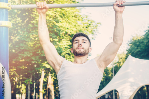 Young bearded muscular male athlete exercising and doing pull-ups on horizontal bar in park early morning, vintage effect